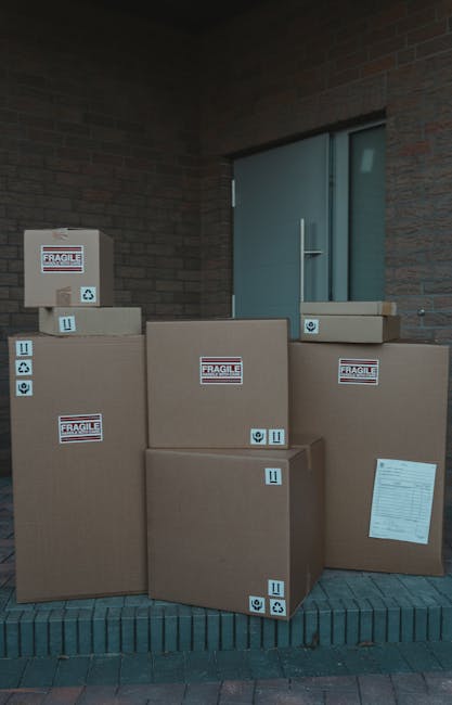 A group of seven large cardboard boxes stacked outdoors on a brick doorstep next to a building with a grey door and brick wall. The boxes vary in size, with some placed directly on the ground and others stacked on top. Each box is sealed with packing tape and features labels indicating fragile contents, along with handling symbols such as arrows and recycling icons. The boxes are prepared for home relocation, likely as part of furniture transport or packing and moving services. The lighting suggests an overcast day, and the scene captures the typical staging of packed items ready for transportation by Man and Van Friern Barnet during a moving process.