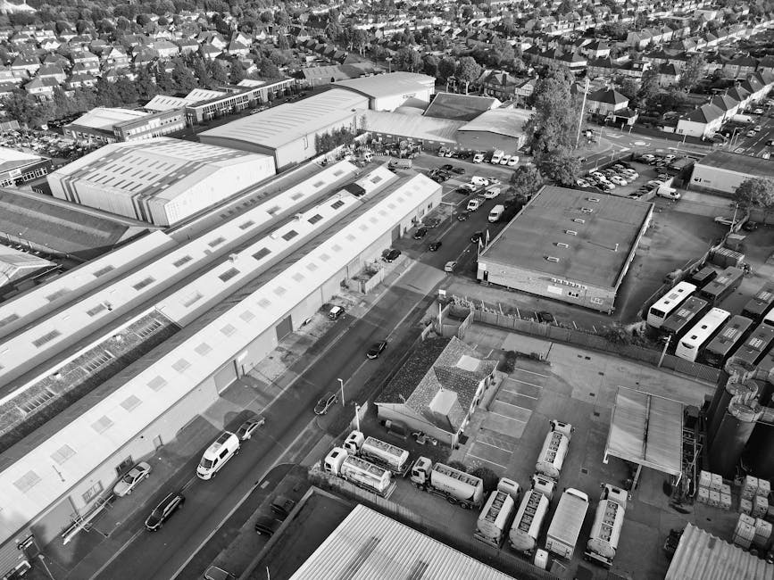 An aerial black-and-white photograph shows an industrial and commercial area with multiple large warehouse buildings, some with flat roofs and skylights, and smaller single-story structures. The scene includes a parking lot with numerous parked vans and cars, a loading area with trucks, and a street with a few vehicles driving through. A small building with a pitched roof is visible near the bottom of the image, surrounded by parked vans and containers. The environment appears to be part of a typical urban logistics hub, with nearby residential neighbourhoods in the distance. This setting illustrates a typical environment for furniture transport and home relocation activities, supported by companies like Man and Van Friern Barnet, who handle packing, loading, and moving services, including loading process and transport logistics.