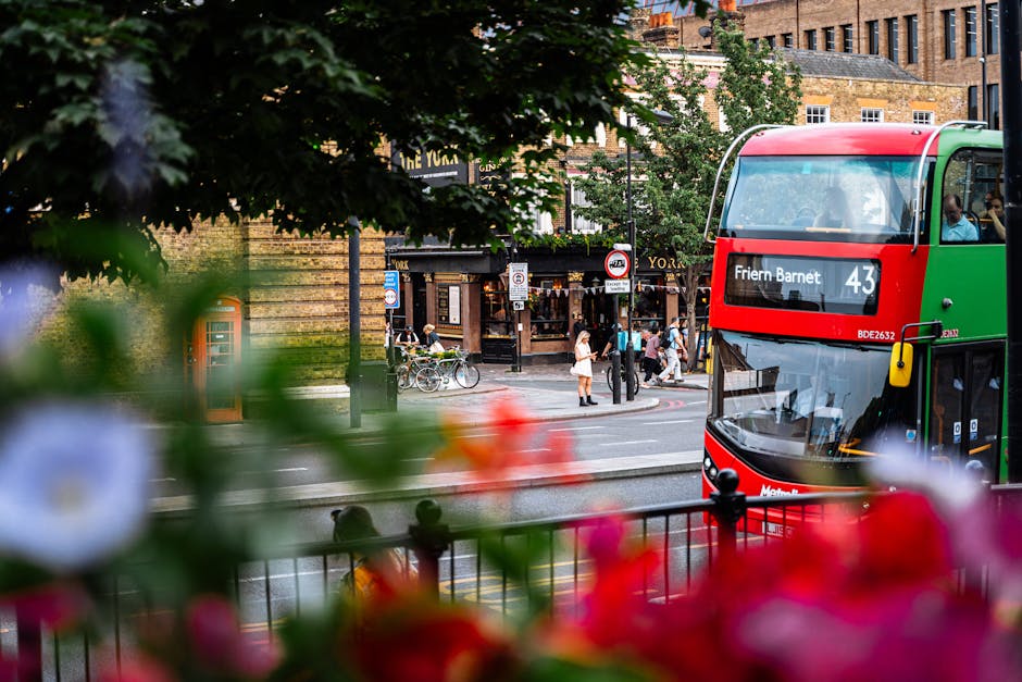 A busy street scene in Friern Barnet with a double-decker bus displaying the route number 43 and the destination Friern Barnet in the foreground. The bus has a green and red exterior and is parked next to a sidewalk. In the background, pedestrians are crossing the street and walking along the pavement, some standing near bicycle racks. There are shopfronts and buildings with brick facades visible across the street, with leafy trees partially obscuring part of the view. Bright flowers in the foreground add color contrast, and an orange public telephone box is situated on the sidewalk. The scene is captured during daylight with natural lighting, illustrating an urban environment suitable for house removals, furniture transport, or home relocation logistics, which are services offered by Man and Van Friern Barnet.