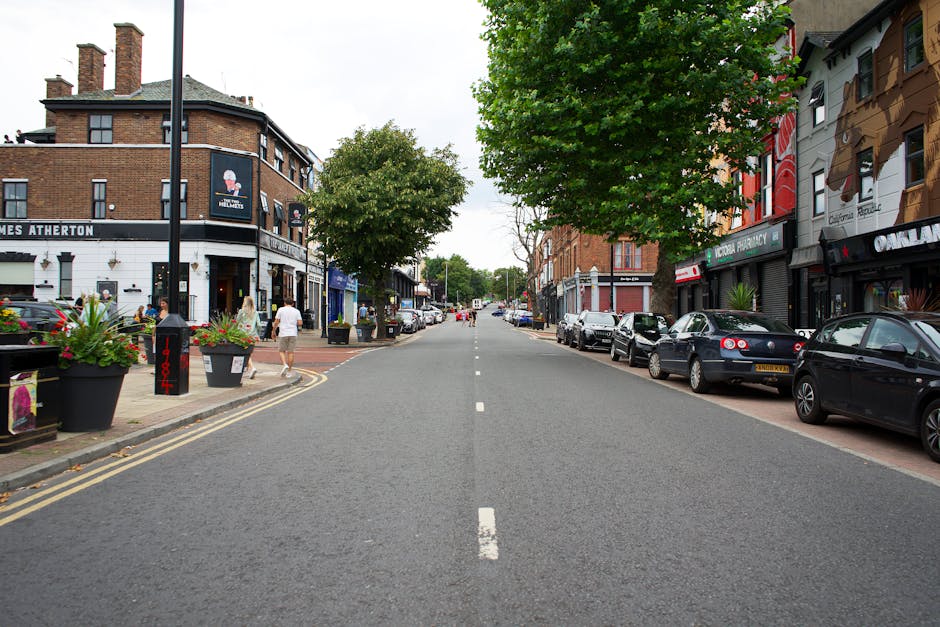 A quiet urban street scene in Friern Barnet during daytime with parked cars lining both sides of the road. On the left, there is a black corner building with a sign for 'James Atherton' and a surrounding pavement decorated with large potted plants featuring colorful flowers. Pedestrians are walking along the sidewalk, including a woman in a white outfit and a man in a dark shirt, as well as other individuals further down the street. The right side showcases a row of shops with closed shutters, including a pharmacy and other retail outlets, with parked vehicles such as black and silver cars adjacent to the pavement. Green trees with full foliage line the street and provide partial shade, while the street itself is empty of moving vehicles and features a dashed white center line. This scene captures the typical setting for local house removals or furniture transport preparations, with an emphasis on parking and accessibility for moving services, as occasionally facilitated by companies like Man and Van Friern Barnet.